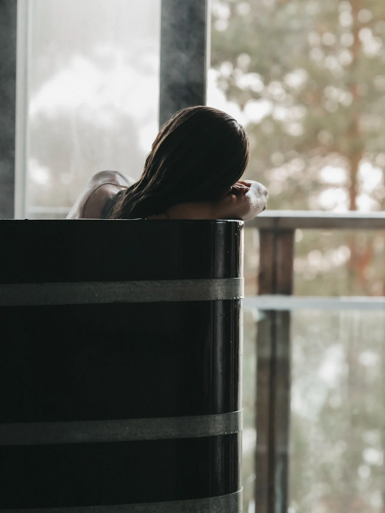 Henkilö rentoutuu höyryävässä kylpytynnyrissä ja katsoo ulos suuresta ikkunasta puihin.