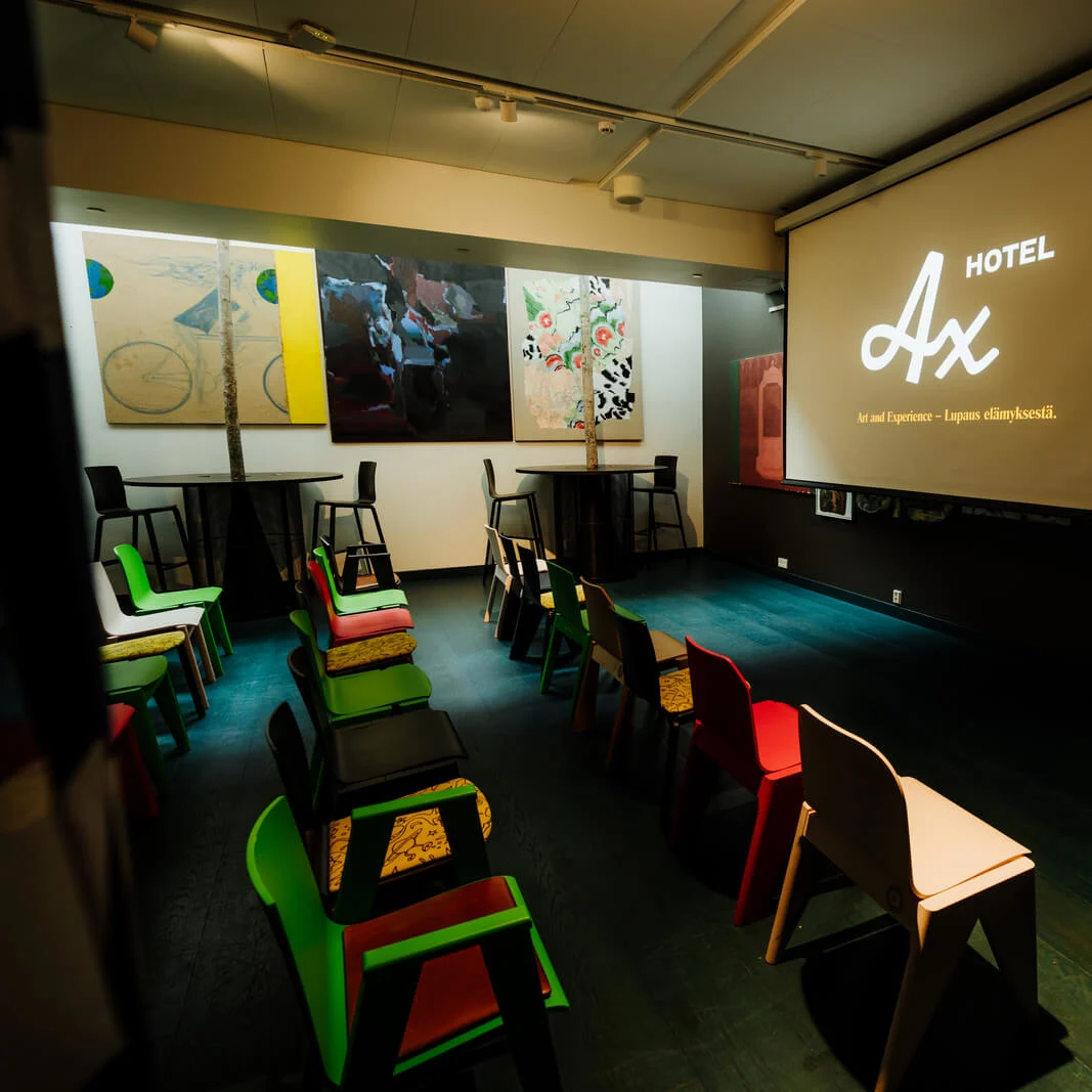 A projection screen shows "Hotel Ax" and "Art and Experience - Lupaus elämyksestä." Colorful chairs face the screen in a dimly lit room with artwork on the walls.