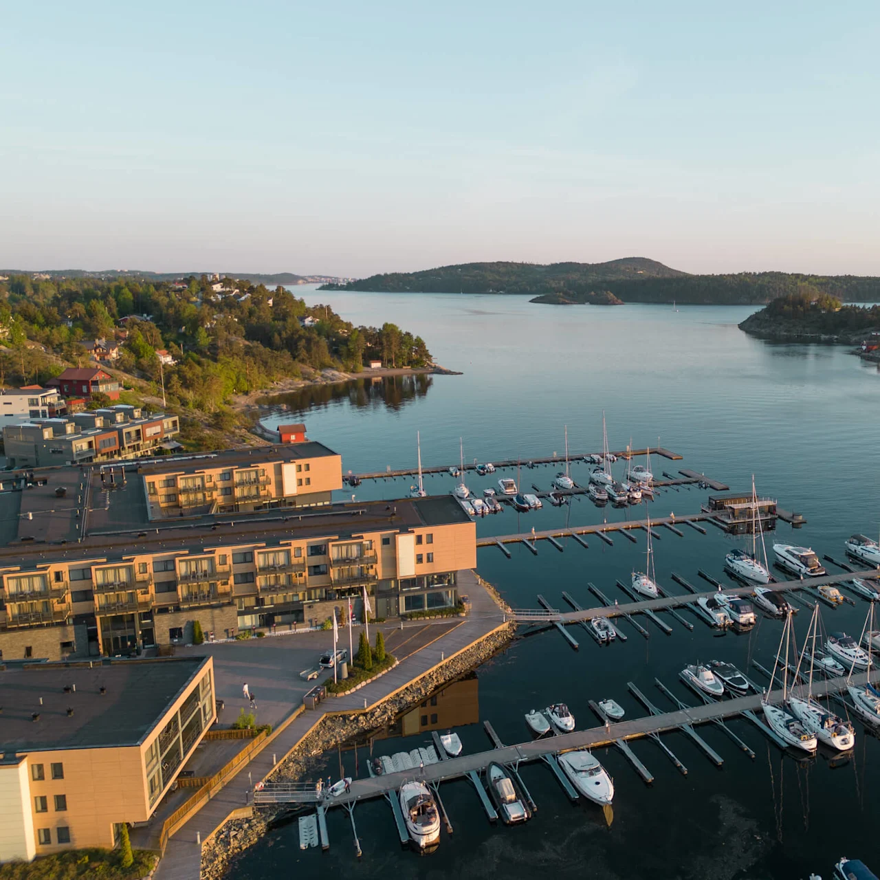 Båter fortøyd i en marina, omgitt av rolig vann. Bygninger langs vannkanten, mot en bakgrunn av frodig grøntområder og små øyer under en klar himmel.