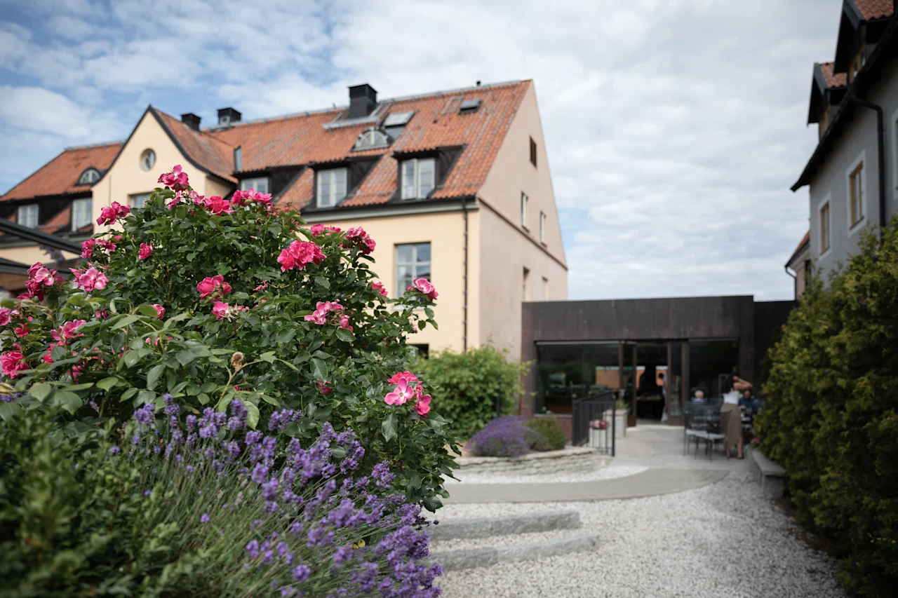 Lyserøde og lilla blomster blomstrer i en have, omgivet af en smal sti. Omgivelserne har en gul bygning med rødt tegltag under en delvist overskyet himmel.