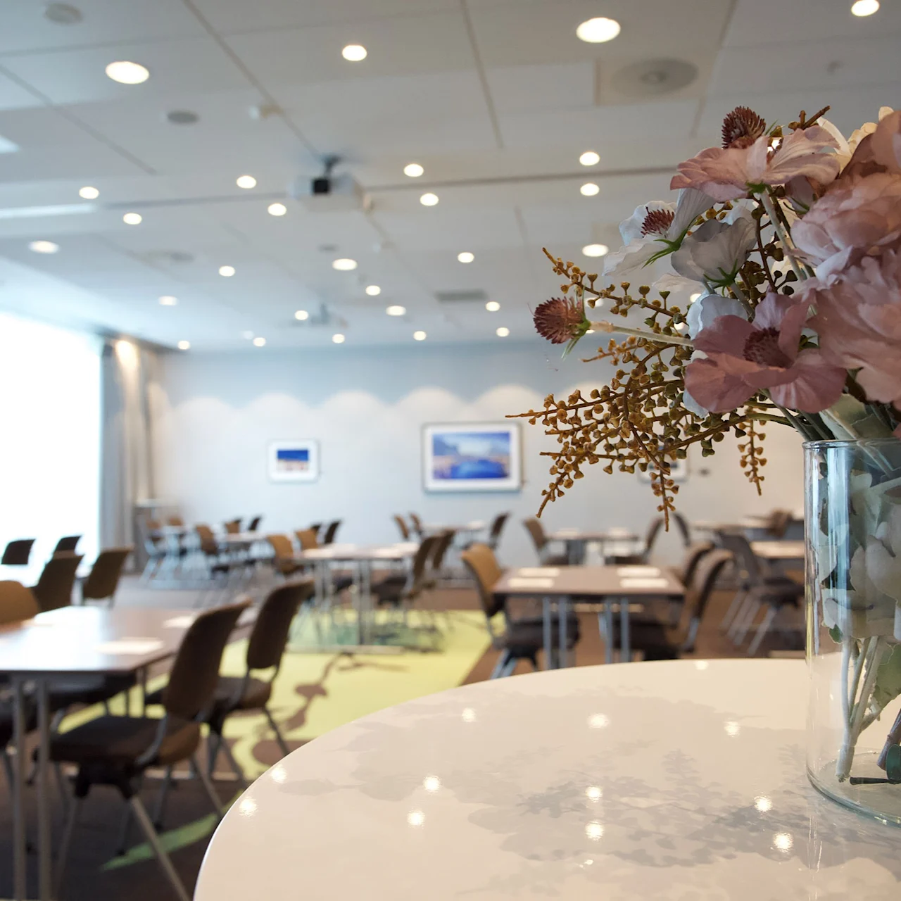 A vase with flowers sits on a round table, overlooking a bright conference room with chairs and tables, under a ceiling with spotlights. Pictures adorn the far wall.