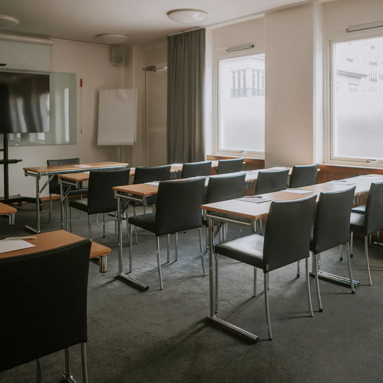 Et konferencelokale med rækker af borde og stole, tomt og pænt, vendende mod en tom whiteboard og et TV.