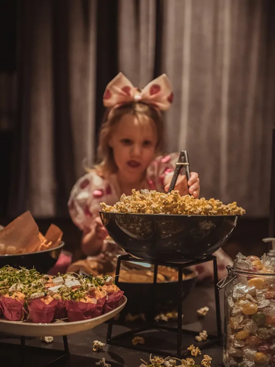 En liten jente med rosa sløyfe i håret forsyner seg med popcorn fra en stor skål på en buffet fylt med muffins, godteri og pizza.