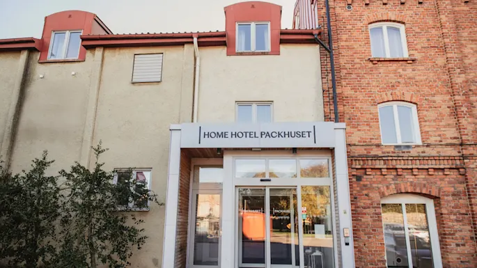 Inngang til en hotellbygning med et skilt som leser HOME HOTEL PACKHUSET over de automatiske glassdørene.