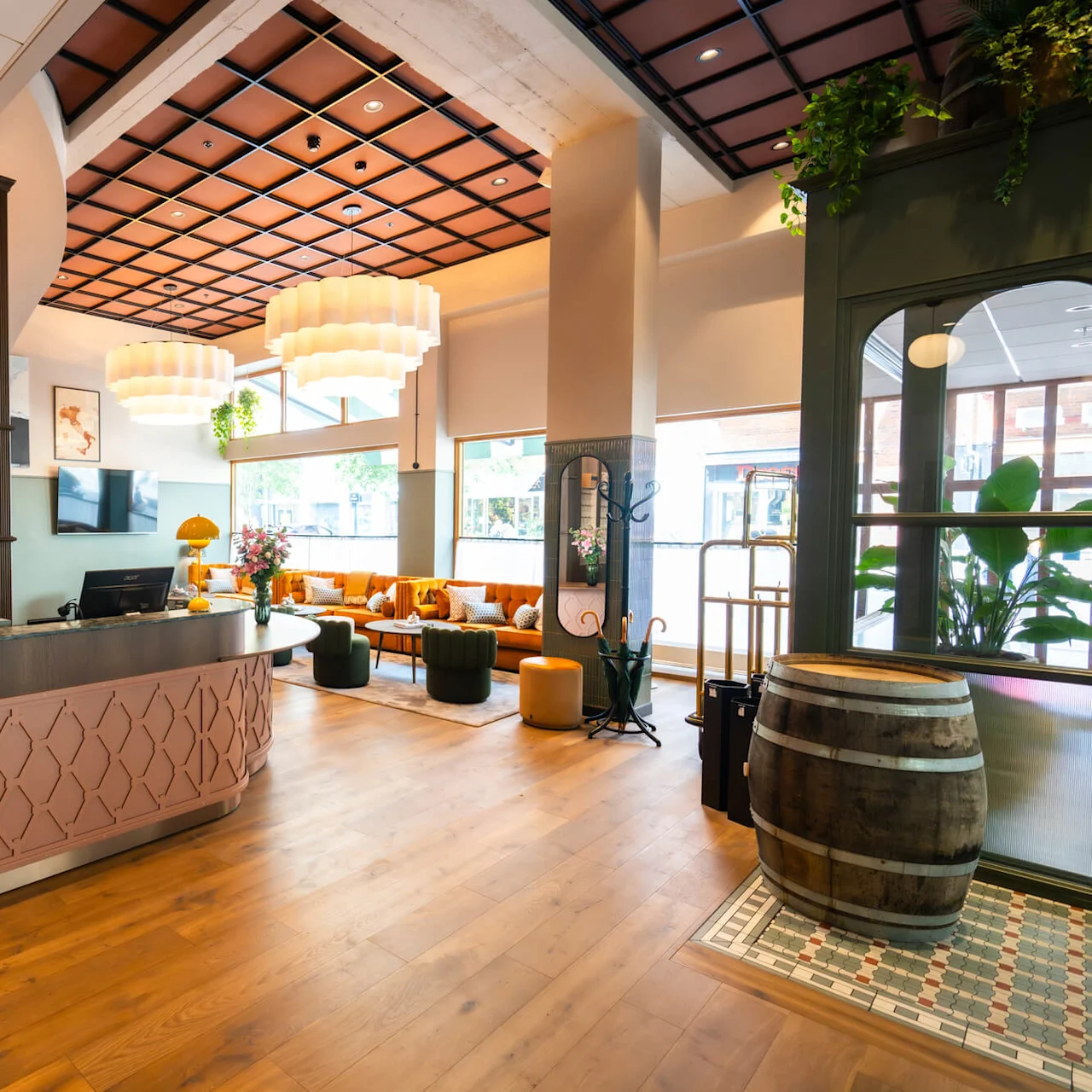 A reception desk with computers and yellow lamps stands in a modern, warmly lit hotel lobby. Orange sofas and decorative plants enhance the inviting atmosphere. 