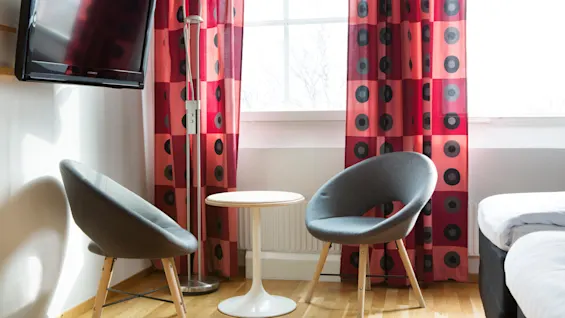 Two gray chairs surround a small white table in front of a window with red curtains featuring black circular patterns. A floor lamp stands nearby, and a TV is mounted on the wall.