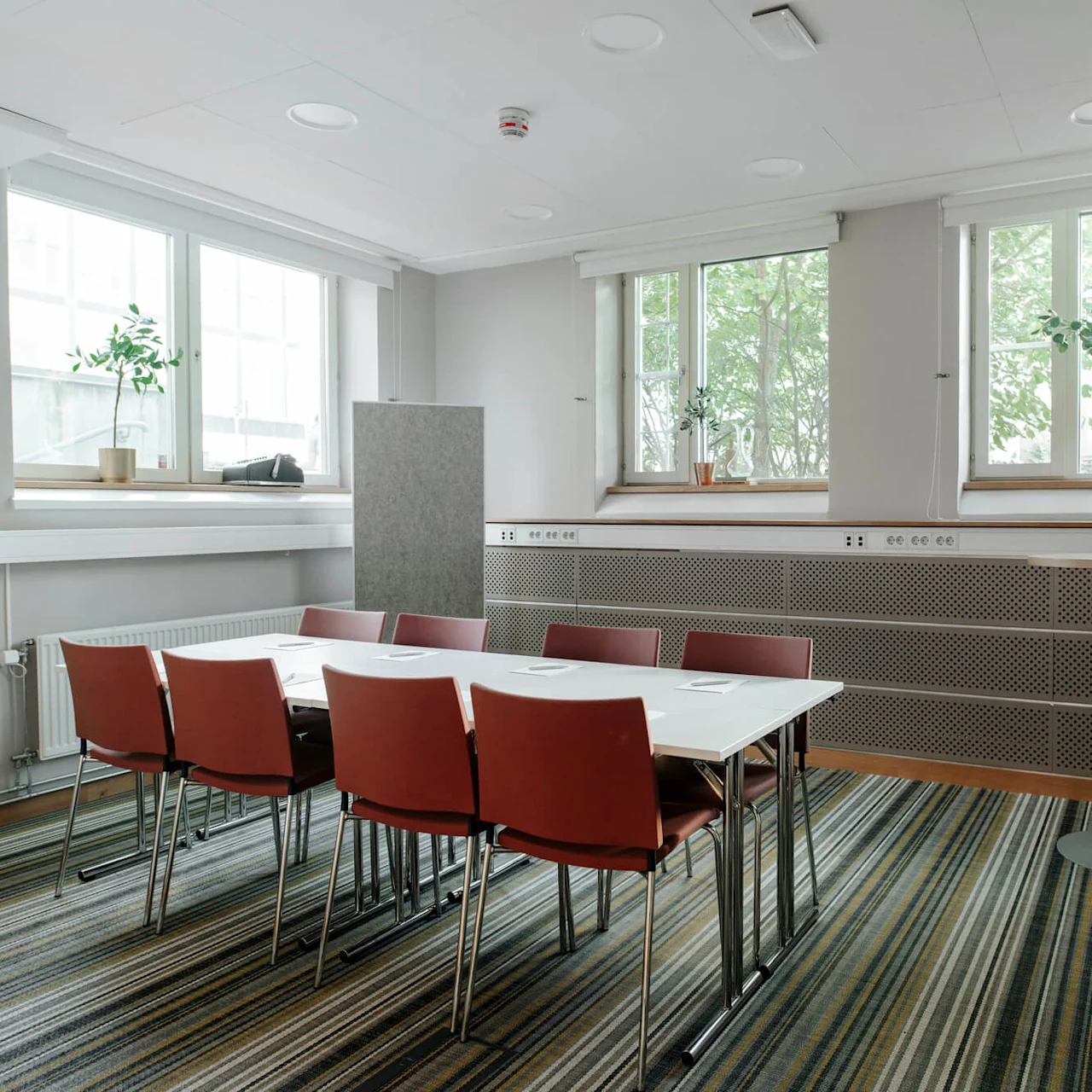 A meeting room with a whiteboard, red chairs, and a table, near a window overlooking greenery.