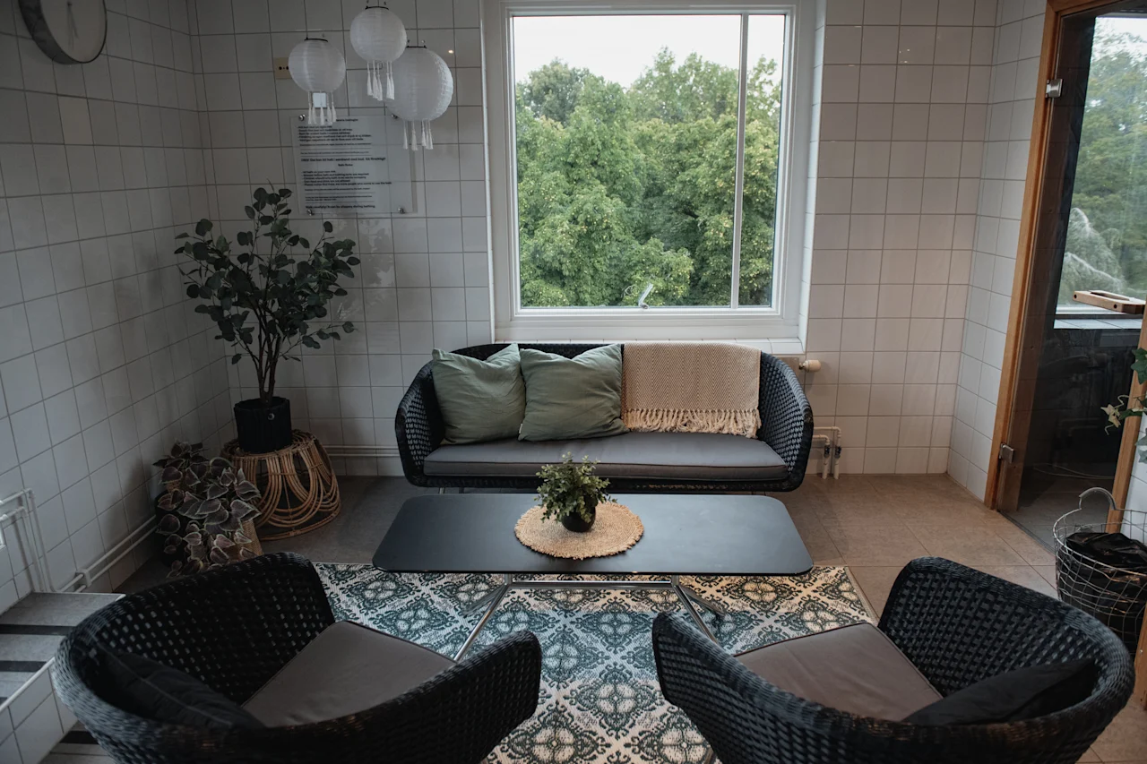 A comfortable lounge area with wicker furniture, green cushions, and a view of lush trees through a large window.