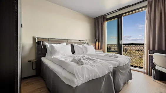 En prydligt bäddad dubbelsäng med vita sängkläder står nära ett stort fönster med utsikt över ett pittoreskt landskap, inramat av bruna gardiner i ett modernt, minimalistiskt sovrum.
