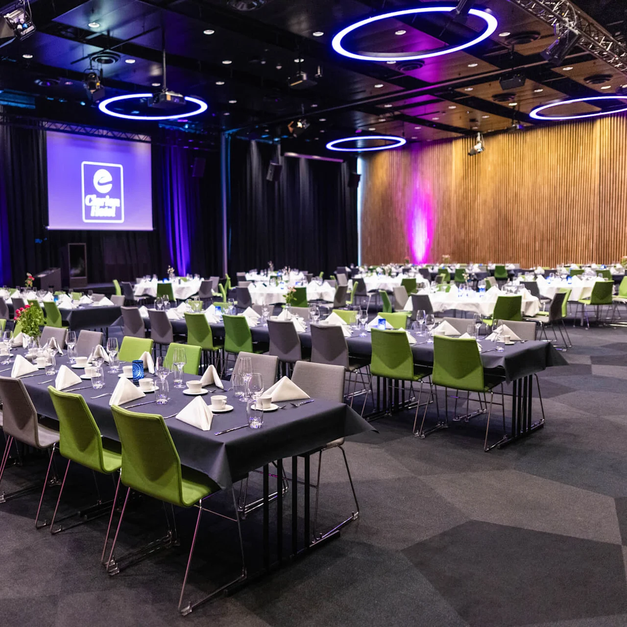 Konferencelokale med rækker af borde dækket med tallerkener, servietter og glas; tomme stole omkranser dem. En stor skærm viser "Clarion Hotel"-logoen. Loftlys kaster et lilla skær.