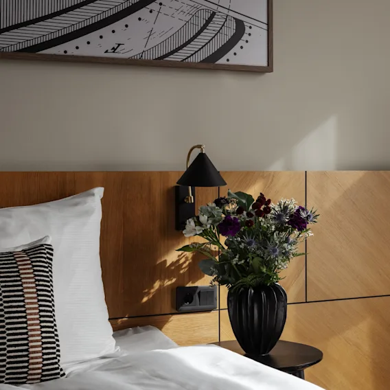 Viihtyisä hotellihuone, jossa sänky, yöpöytä kukkamaljakolla ja abstrakti taideteos seinällä.
