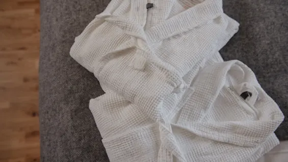 Two white waffle-patterned bathrobes lie folded on a gray fabric surface, beside a wooden floor. 