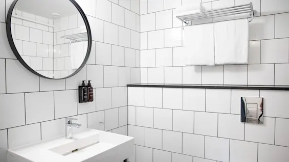 Et badeværelse med hvide fliser har en rektangulær håndvask med en moderne vandhane, et rundt spejl over, og hylder med foldede håndklæder. Shampoo flasker er fastgjort til væggen ved spejlet.