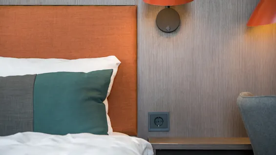 Bed with orange headboard and green-brown pillow resting in a modern room. Lit orange lamp hangs on the textured wall above a power outlet.