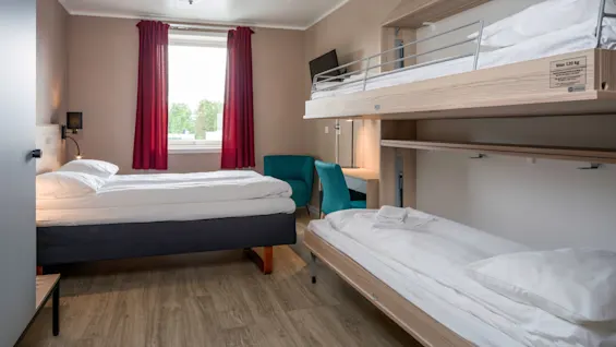 A tidy hotel room features a large bed and bunk beds beneath a small desk, accented by red curtains near a window overlooking trees. A sign reads "Max 120 kg."
