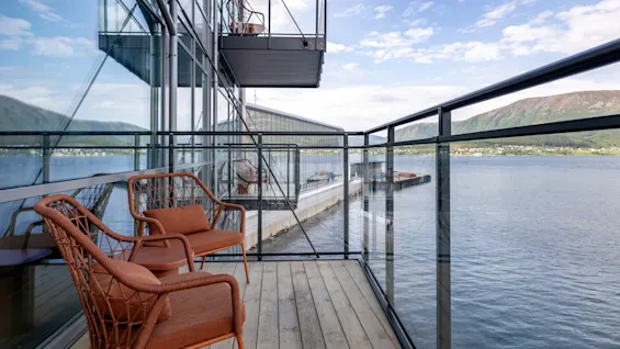 To orange stole står på en træbalkon med udsigt over en sø. Omgivet af et glasrækværk omfatter udsigten fjerne bjerge under en delvis overskyet himmel.