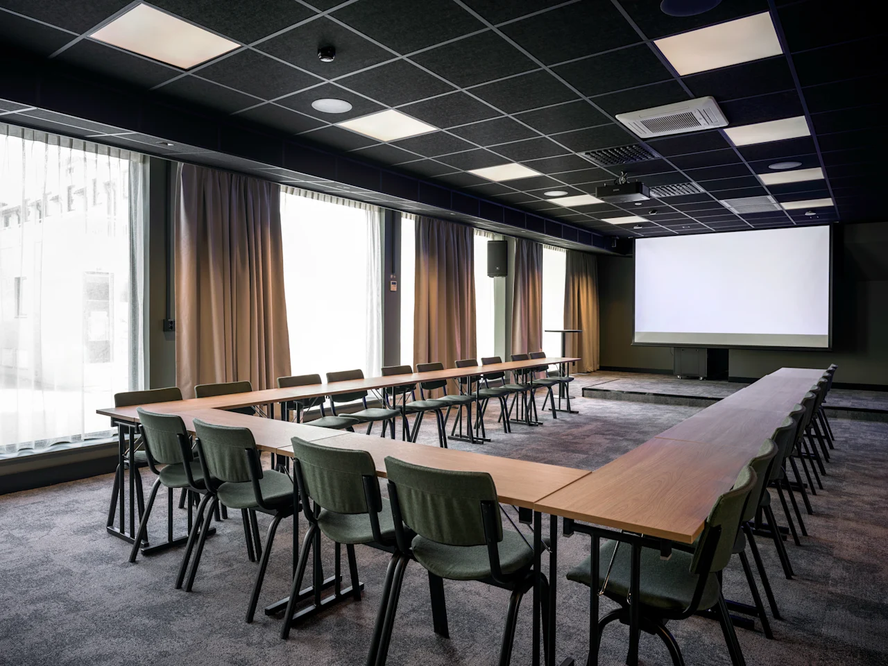 A modern conference room with tables and chairs arranged in a U-shape, facing a large projection screen. Large windows line one wall.