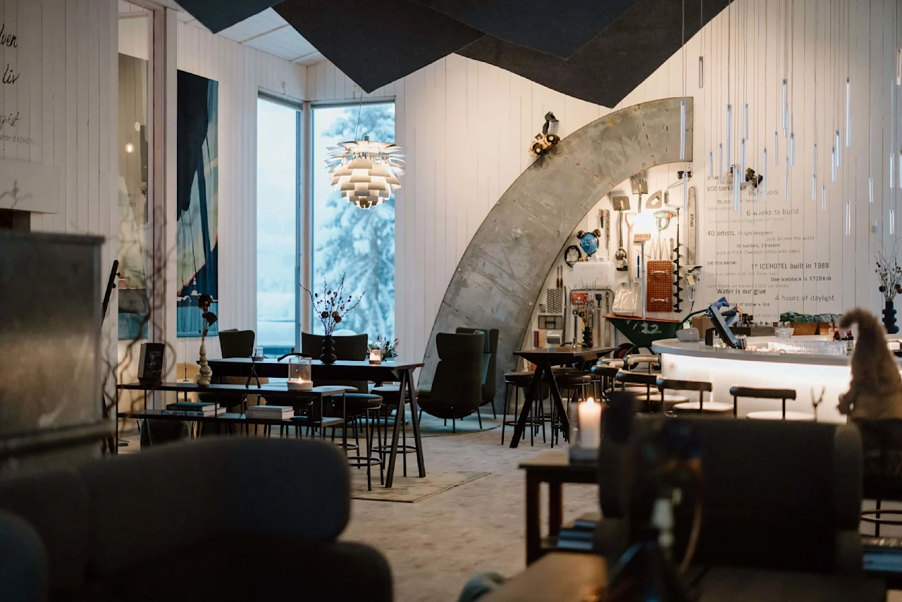 Et hyggeligt rum med moderne møbler og dekorativ belysning. Et vindue viser snedækkede træer udenfor. Tekst på væggen inkluderer: "ICEHOTEL built in 1989," "600 tons of ice," "12 weeks to build."