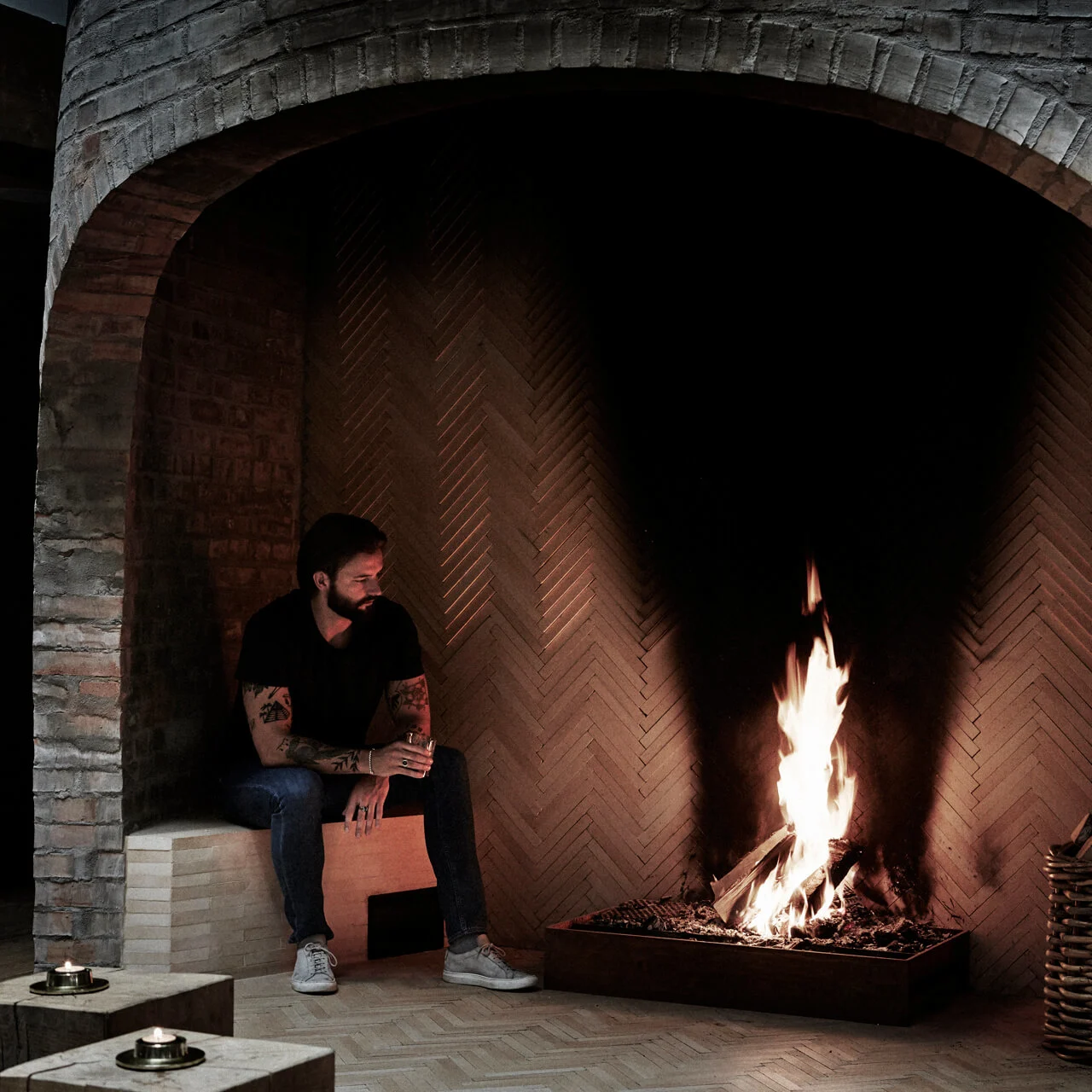 Person sits beside a large brick fireplace, holding a drink, watching the flames. A basket filled with logs is nearby in the dimly lit, cozy room.