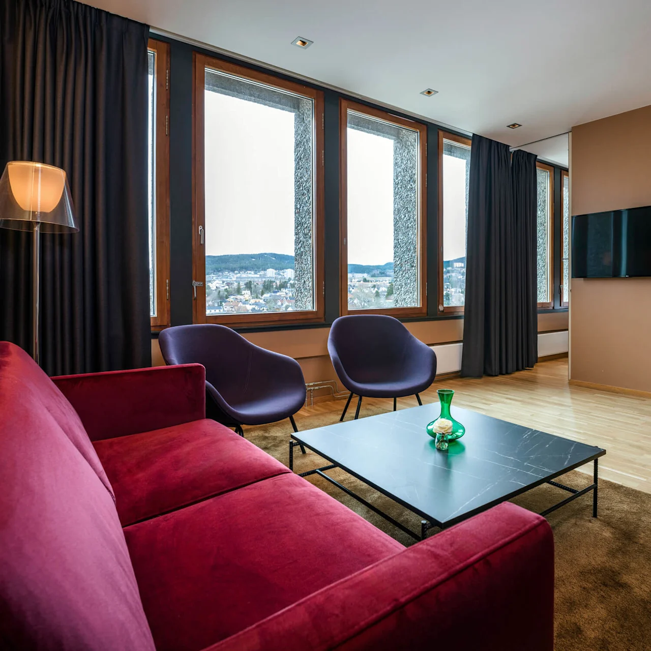 Red sofa and two purple chairs face a low black table in a modern, cozy living room with wooden floors. Large windows offer a view of a cityscape and mountains.