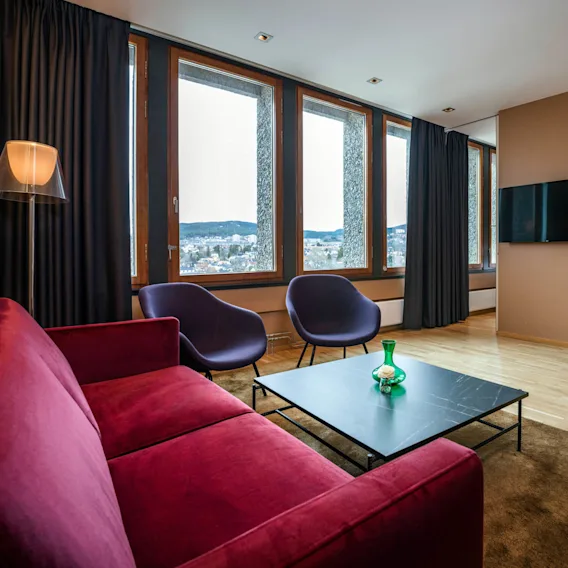 Rød sofa og to lilla stoler vendt mot et lavt svart bord i et moderne, koselig stue med tregulv. Store vinduer gir utsikt over bybildet og fjellene.