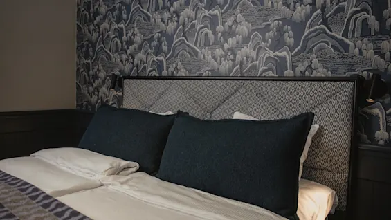 Ett mysigt sovrum med en säng med mönstrad sänggavel och mörka kuddar, mot en vägg med intrikat blå och vit tapet.