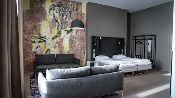 A modern room features a gray sofa facing a colorful wall mural labeled "The Sacrifice," with adjacent neatly made twin beds. A hanger and mirror are on the right side.