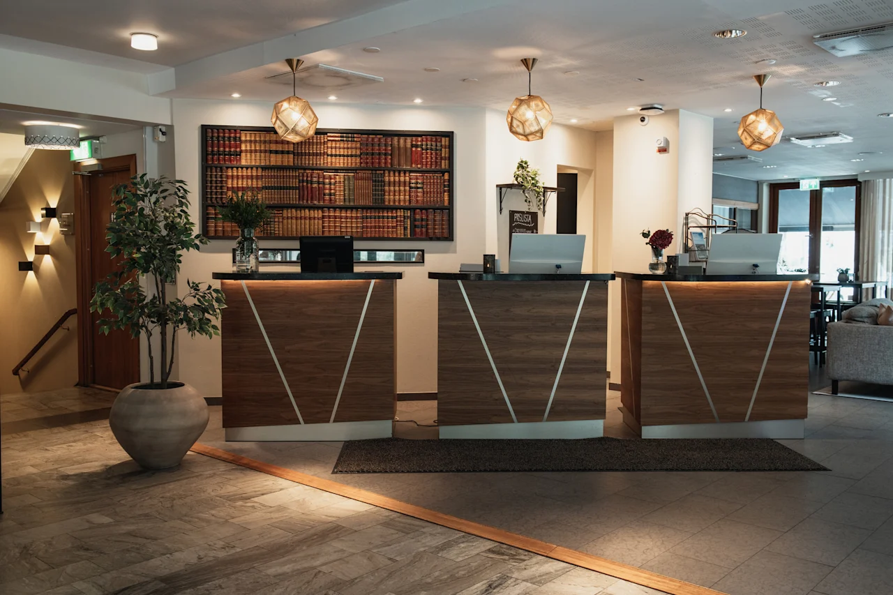 A stylish hotel reception area with three service desks, computers, and a decorative wall of books. A sign reads PRELUSA.