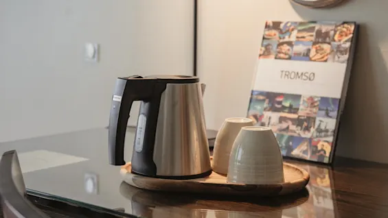 En vattenkokare och två koppar på en bricka, med en bok med titeln TROMSO i bakgrunden, vilket antyder en mysig dryckesuppsättning.