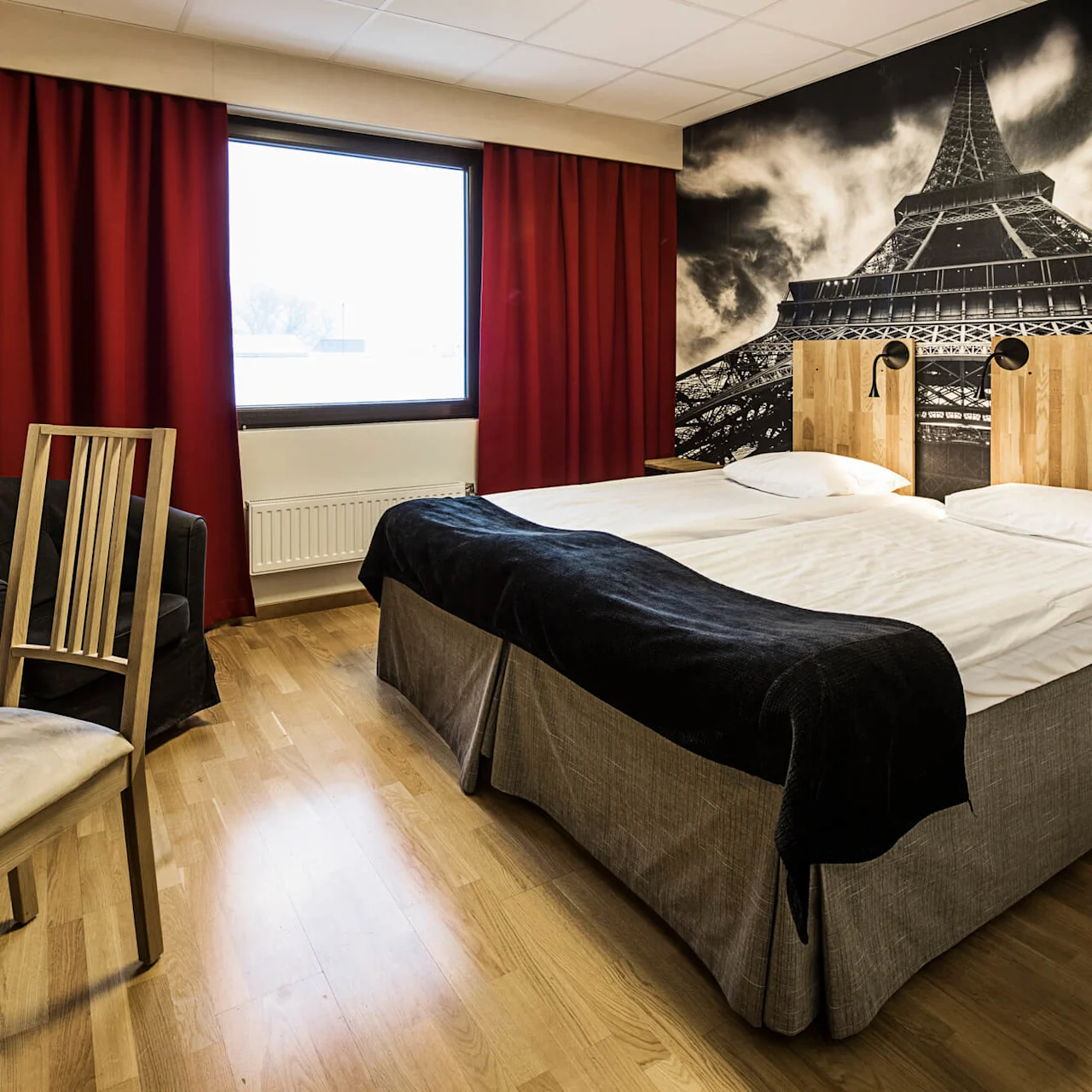 A hotel room with twin beds, dark red curtains, a large mural of the Eiffel Tower, and simple furniture, suggesting comfort and modern interiors.