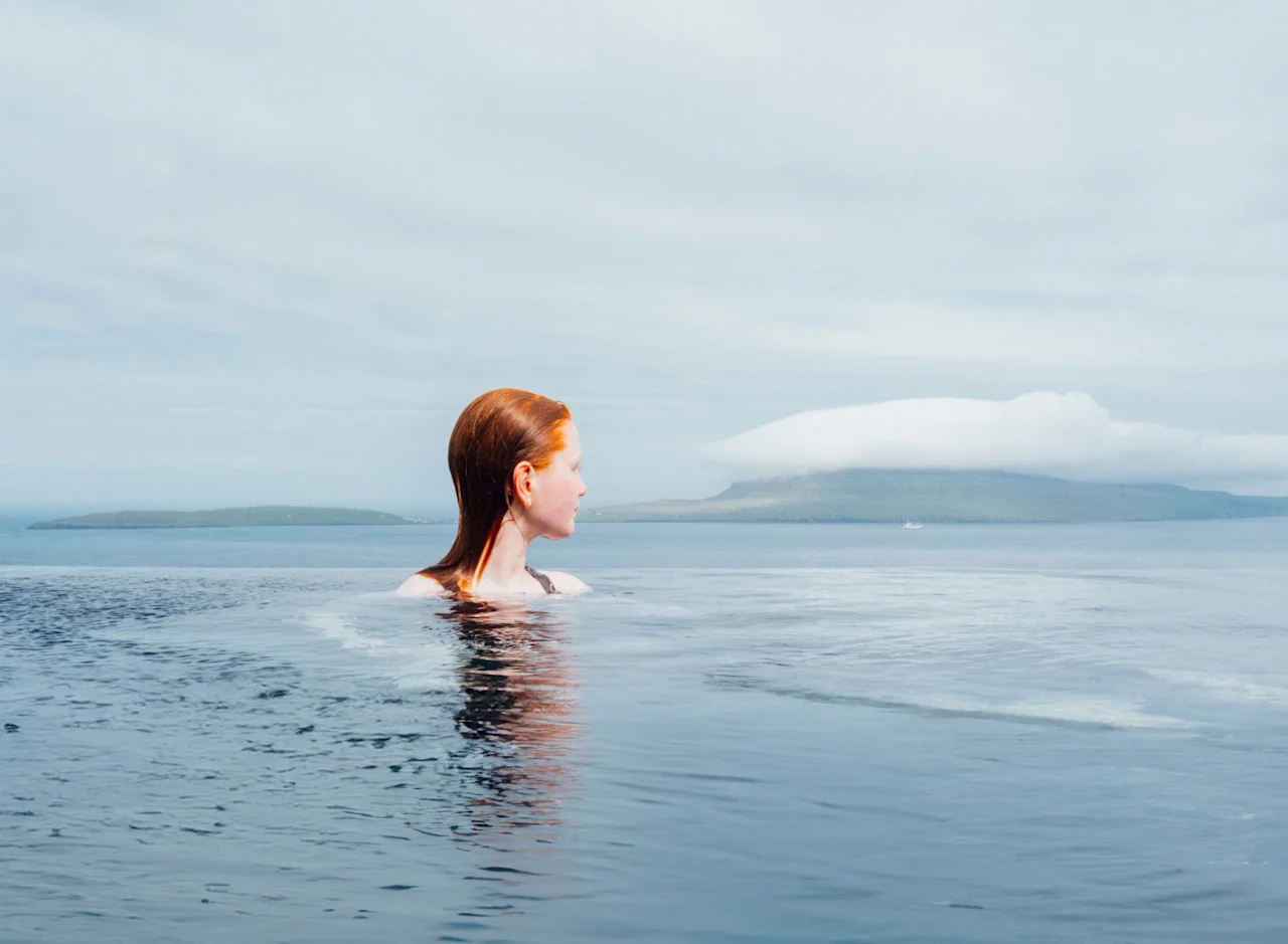 En person med rött hår i en infinitypool, som blickar ut över ett lugnt havslandskap med avlägsna öar under en molnig himmel.