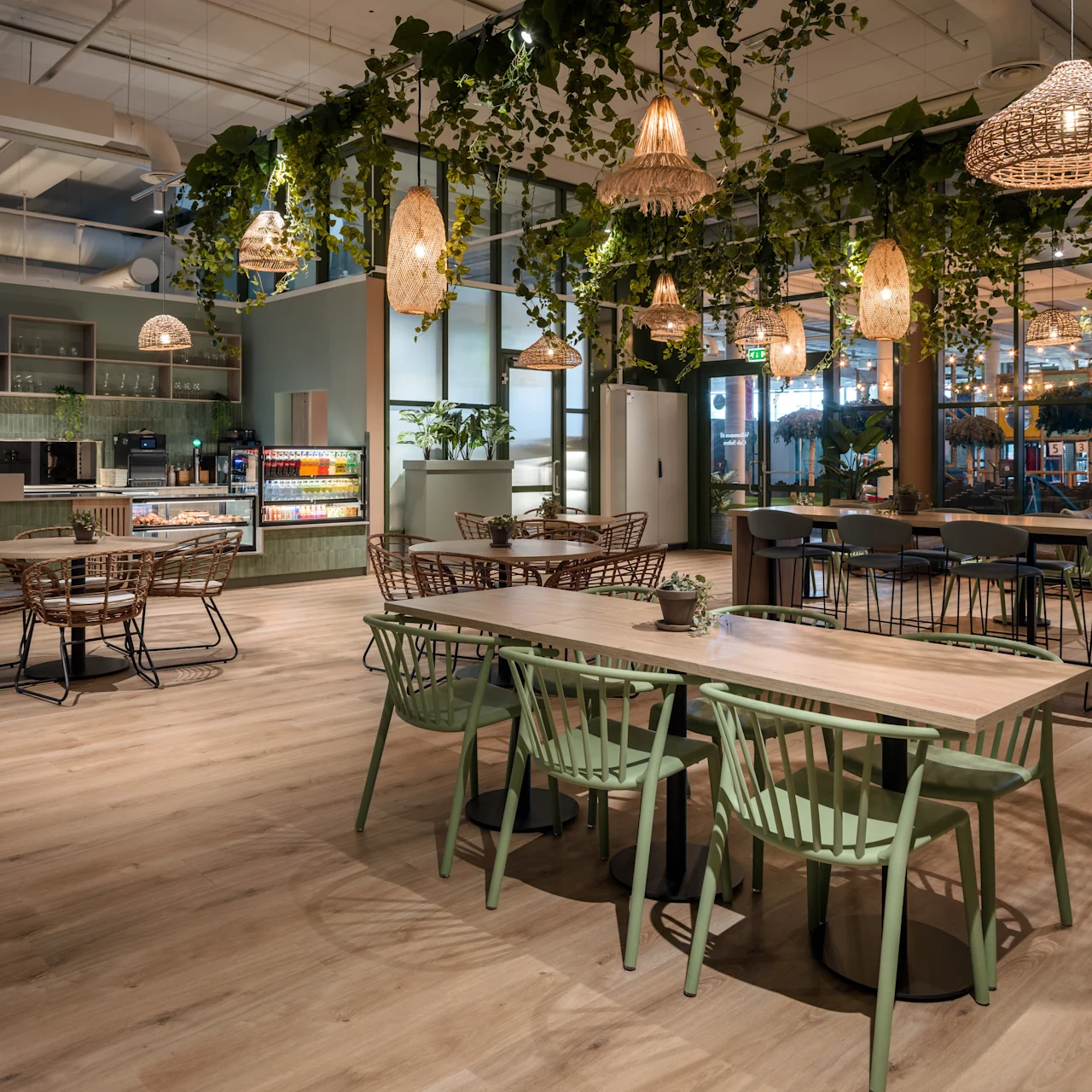 Tables and chairs are arranged in a modern café, with hanging lights and greenery above, within a spacious, well-lit interior featuring wooden floors and shelves filled with kitchen items.