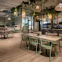 Borde og stoler er arrangert i en moderne kafé, med hengende lys og grønt over, i et romslig, godt opplyst interiør med tregulv og hyller fylt med kjøkkenartikler.