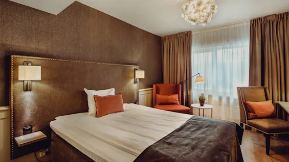 A neatly made bed with an upholstered headboard in a well-lit, luxurious bedroom with armchairs, curtains, and a chandelier.