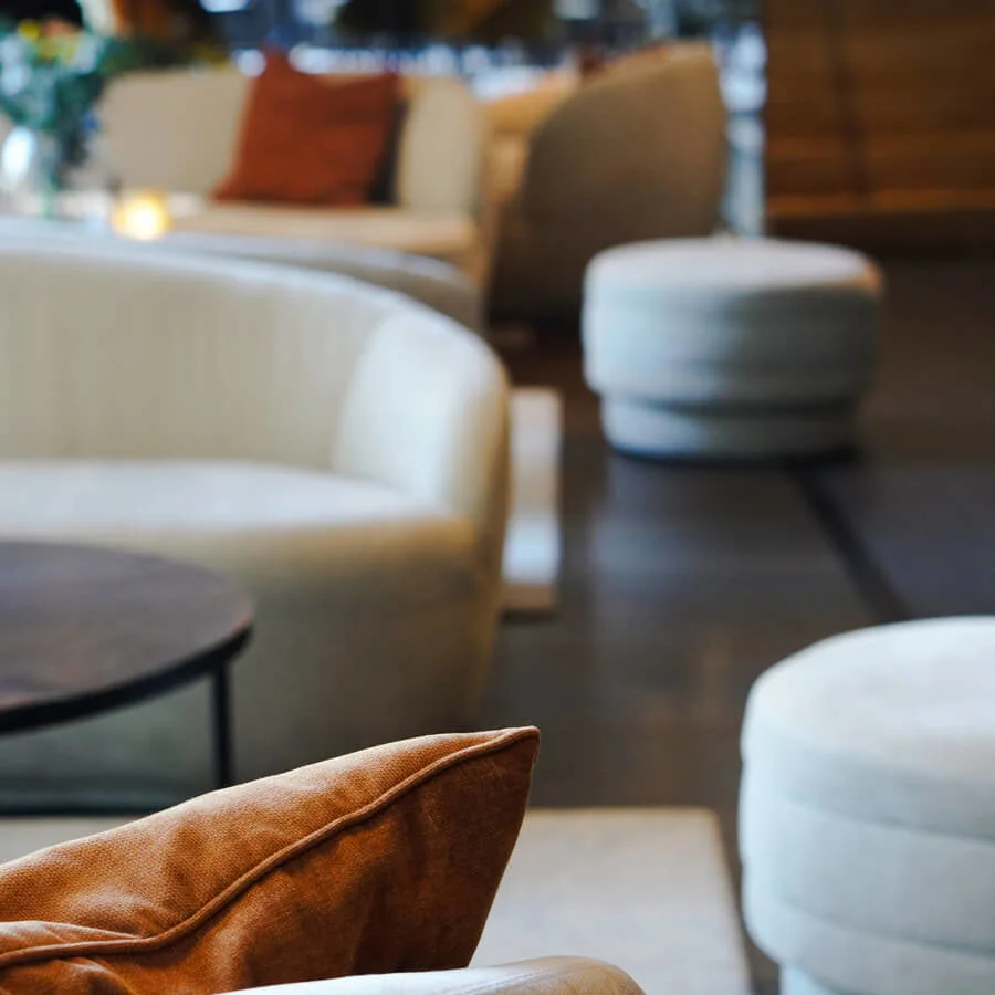 The room features round, beige sofas with plush cushions, arranged around low coffee tables. Soft lighting and blurred background emphasize a cozy, modern lounge atmosphere.