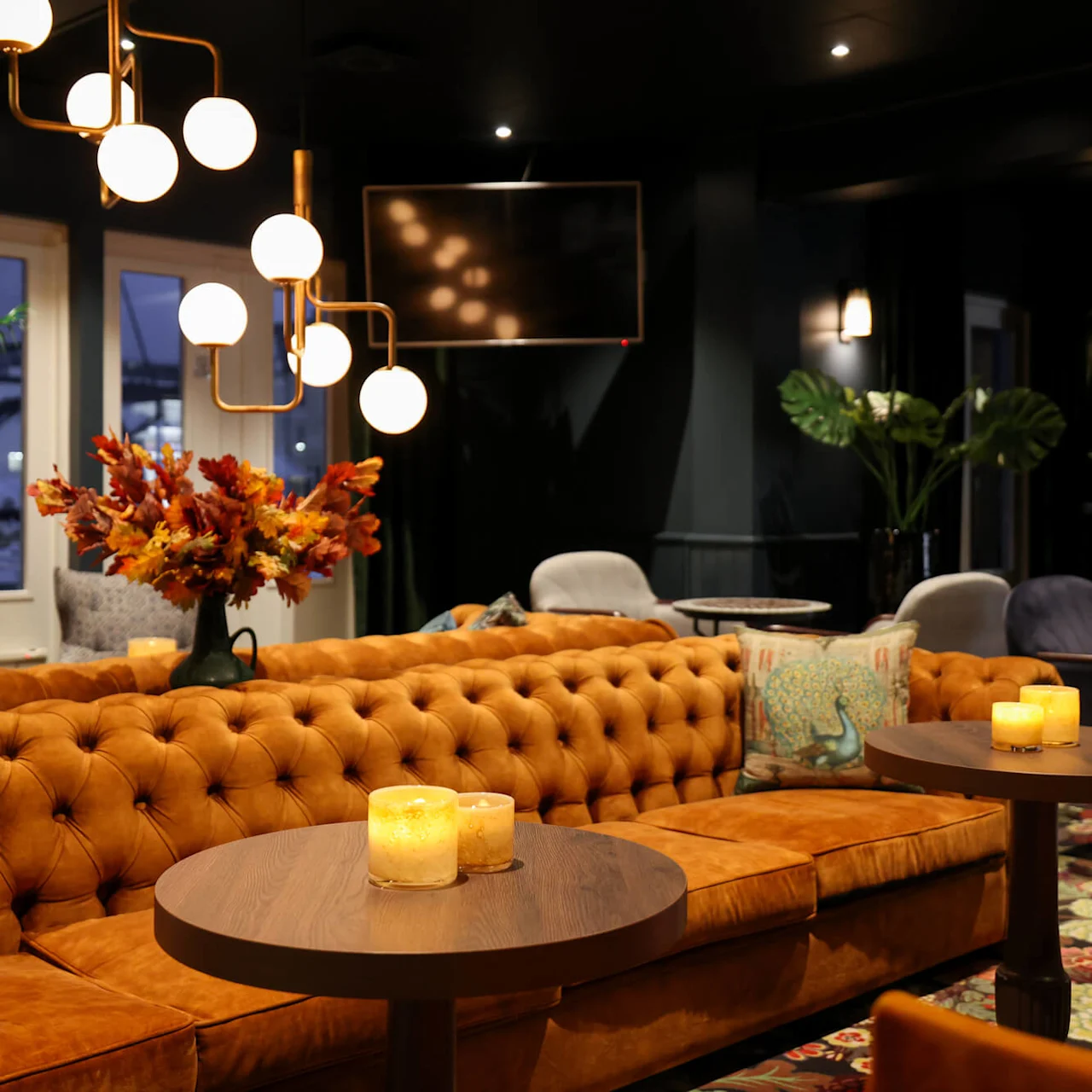 A tufted orange sofa with lit candles on round tables, set in a dimly lit, elegant lounge featuring modern spherical chandeliers, decorative pillows, and plants near large windows.