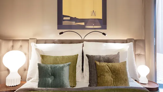 A neatly made bed features multiple textured pillows and a green blanket, flanked by modern lamps. Above, a minimalist artwork hangs on a beige wall in a softly-lit bedroom.