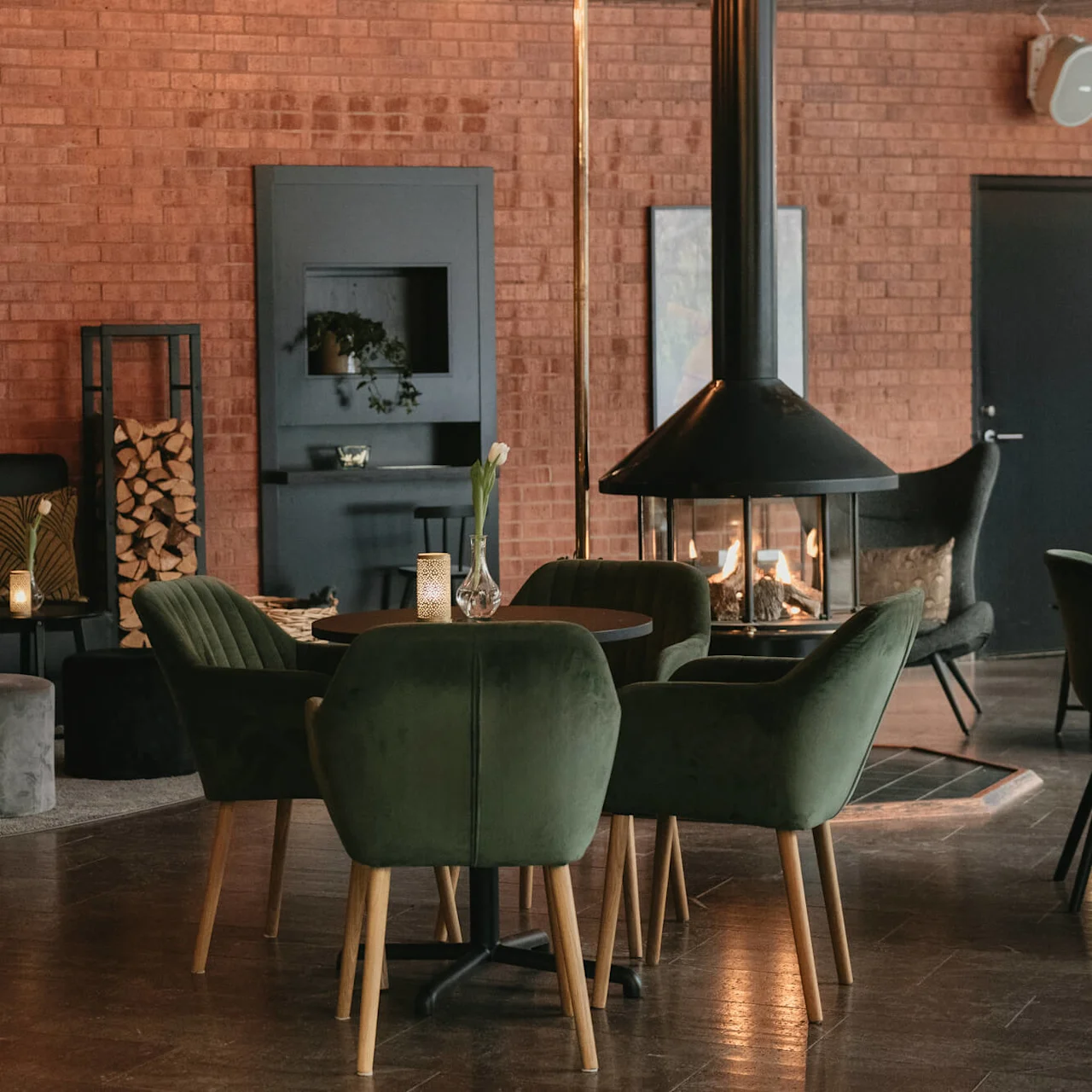 Cozy lounge with green velvet chairs surrounding a table. A nearby fire flickers in a modern fireplace. Brick walls enhance the warm, inviting atmosphere with mirrors and plants for decoration.