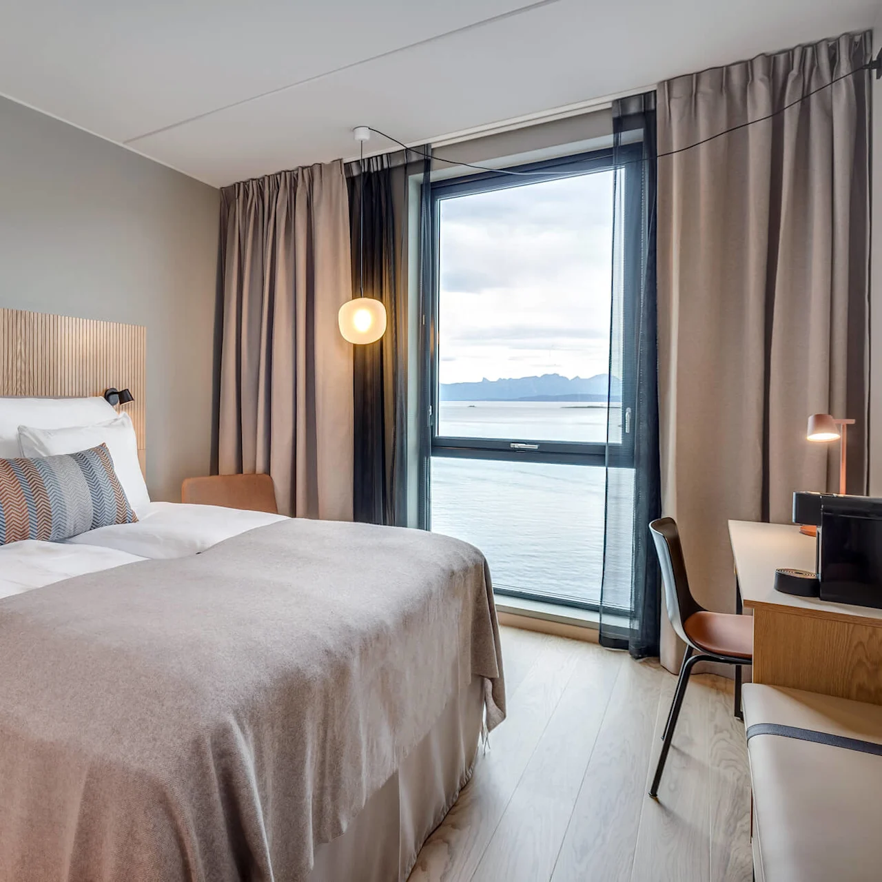 Et hyggeligt hotelværelse med en redt seng, moderne lampe, skrivebord og et stort vindue med udsigt over havet og fjerne bjerge.