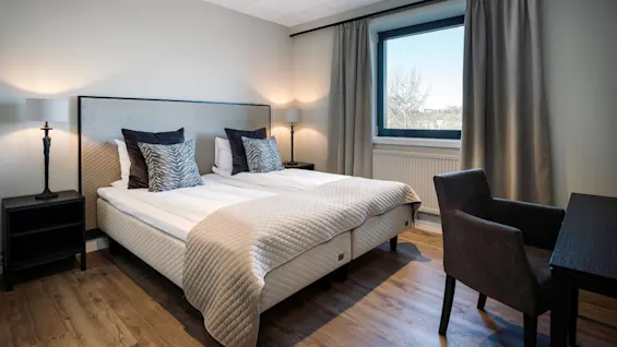 Seng med mønstrede puder står i et pænt rum. To lamper på sideborde oplyser rummet, mens et vindue viser udsigten. En stol vender mod et skrivebord.