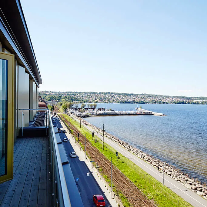 Bygning med åben altandør har udsigt over kystlinjen. Jernbanespor og vej løber parallelt ved siden af et græsareal, mod en fjern by langs vandkanten under en klar himmel.