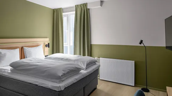 En snyggt bäddad säng med vita lakan står mot en olivgrön accentvägg, bredvid ett fönster med gröna gardiner. En svart golvlampa står bredvid ett element.