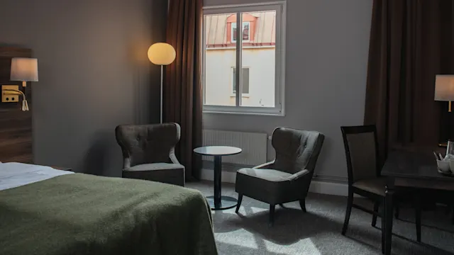 Cozy chairs sit beside a small round table under a soft-lit lamp in a hotel room. A bed with a green cover and a window view complete the tranquil setting.