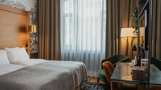 Et hyggeligt hotelværelse med en seng, et skrivebordsområde og et vindue med gardiner, der antyder et behageligt ophold.