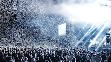 En stor folkemengde på en konsert med sterke scenelys og konfetti som faller, noe som skaper en energisk atmosfære.