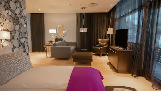 Et hotellrom med en dobbeltseng har et lilla teppe. I nærheten står en elegant sofa og lenestol på lyst tregulv, med en TV og gulvlange gardiner.