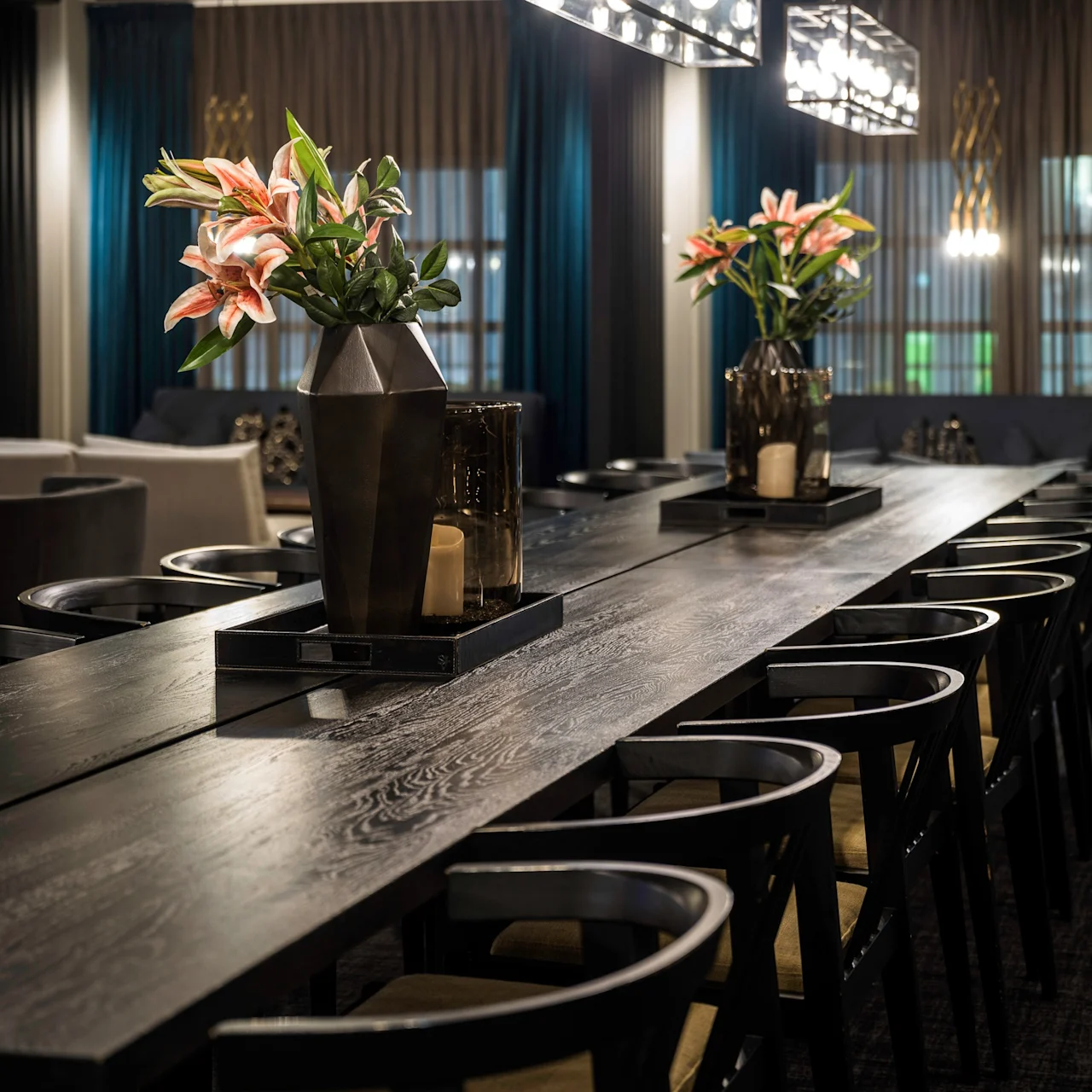 Long wooden dining table holds decorative vases with pink lilies, surrounded by black chairs. Modern pendant lights hang above, set in a dimly lit, elegantly furnished room.