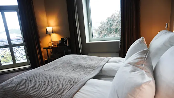 En seng med hvide puder og et gråt tekstilteppet hviler i et hyggeligt rum. En lampe lyser varmt ved siden af et vindue med udsigt.