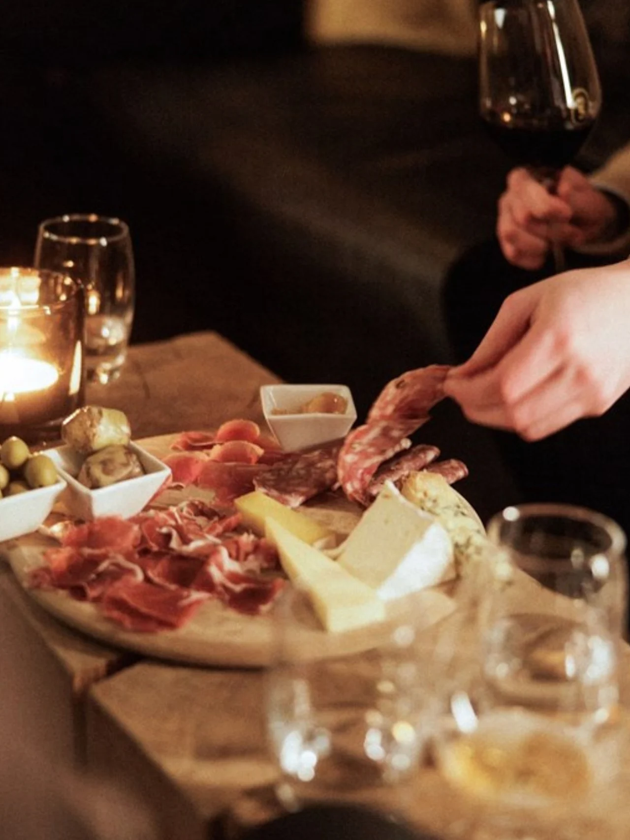 A charcuterie board with meats, cheeses, and olives on a wooden table; a hand reaches for salami slices, while another holds a glass of red wine. Dimly lit ambiance.