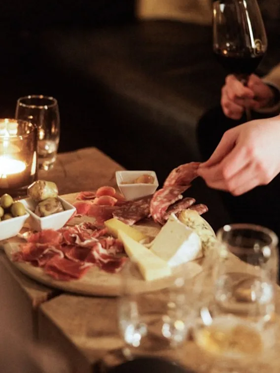 A charcuterie board with meats, cheeses, and olives on a wooden table; a hand reaches for salami slices, while another holds a glass of red wine. Dimly lit ambiance.